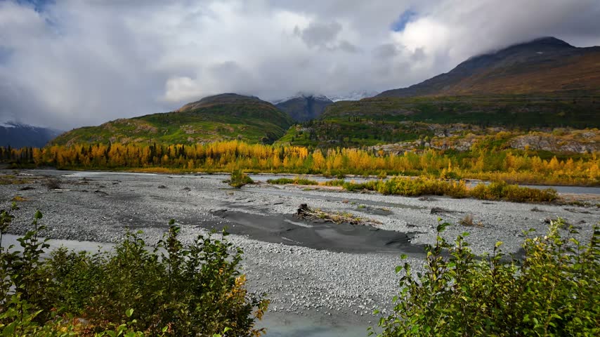 Moving clouds casting shadows over a vast riverbed and mountains in the Alaskan wilderness.