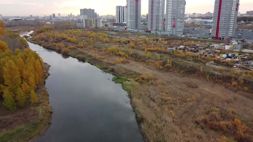 Aerial view of river flowing through autumn landscape. Yellow trees line riverbanks in golden light. Modern buildings rise behind the natural scenery. River curves gently, reflecting twilight sky