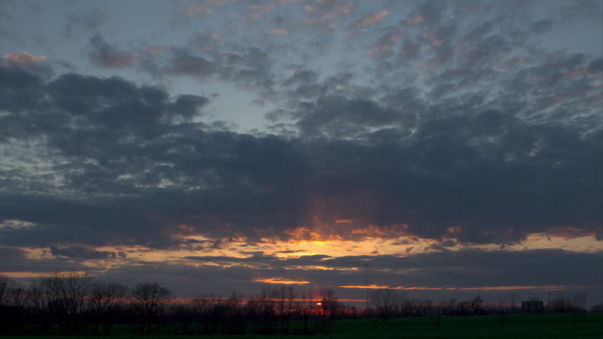 Red sun setting below the horizon in the American countryside. Dark clouds move across the dramatic twilight sky over a green field
