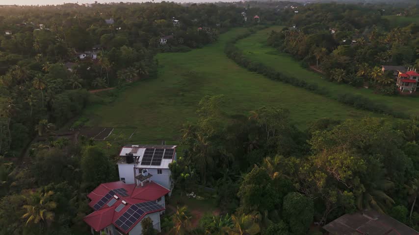 Drone landscape at sunrise showing tropical village rooftops and palm trees beside wide green fields and misty countryside, peaceful rural morning scenery.