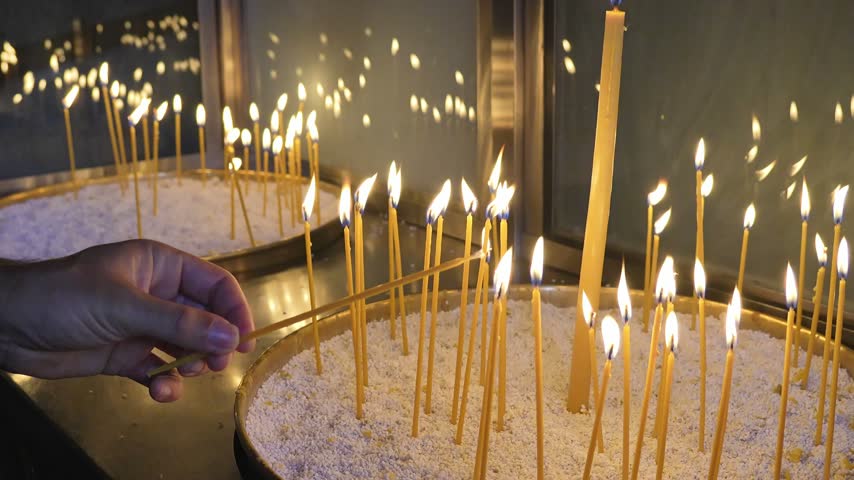 Hands performing a religious ritual, lighting a thin candle among many flickering flames in a container of sand in an Orthodox church in Greece