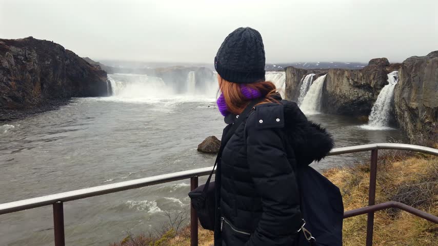 Back view of a female traveler in a winter jacket and wool hat standing at the viewpoint railing, gazing at the thundering horseshoe of Godafoss in north Iceland.