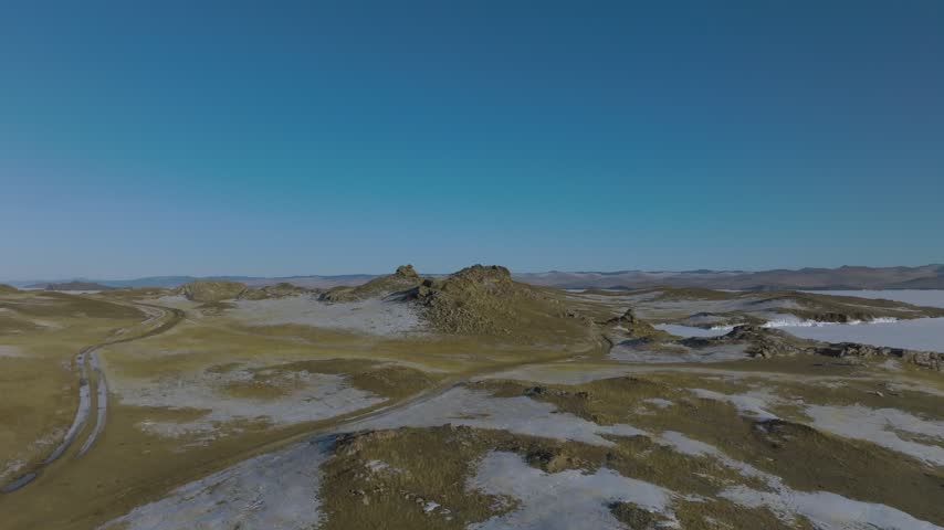 Wide scenic view showing the frozen expanse of Lake Baikal surrounded by snow covered rocky hills and distant mountains in Irkutsk Oblast, Russia under a clear bright blue winter sky
