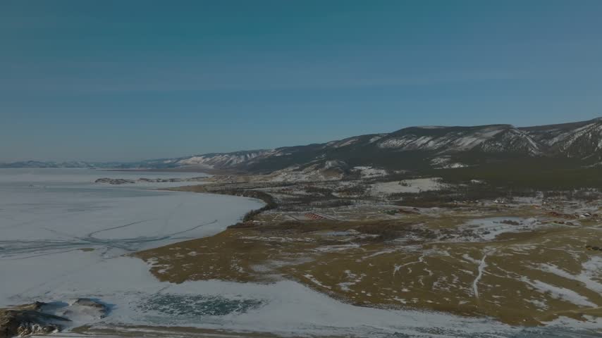 High angle aerial view over the frozen ice sheet of Lake Baikal along the shoreline of Irkutsk Oblast, Russia, showing snowy hills, tire tracks, and endless winter landscape