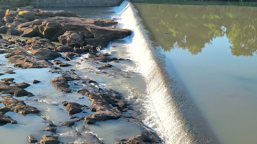 A tranquil scene of a small waterfall flowing over a stone weir on a calm river with reflections and natural rocks in the foreground. 4K