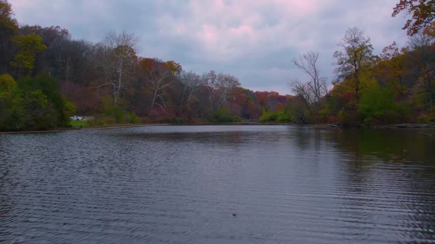 Drone view over a calm lake surrounded by colorful fall woodland beneath a cloudy sky at Mill Creek Park in Youngstown, Ohio.
