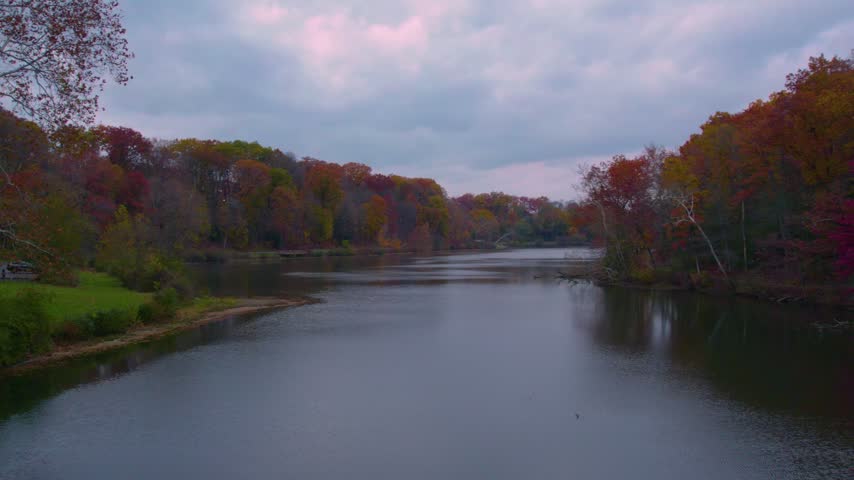 Drone view across a broad lake surrounded by colorful autumn forest at Mill Creek Park in Youngstown, Ohio under a soft cloudy sky.