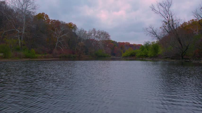 Low aerial drone view skimming above calm lake water surrounded by autumn trees at Mill Creek Park in Youngstown, Ohio.