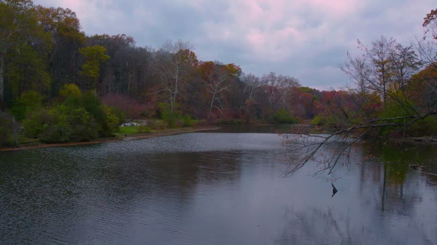 Drone view across a peaceful lake surrounded by colorful autumn woodland and a cloudy sky at Mill Creek Park in Youngstown, Ohio.