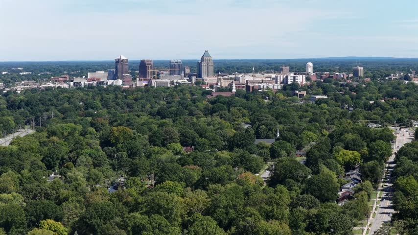 Downtown towers and a white water tower stand on the horizon above a wide green canopy and a road running between leafy neighborhoods in Greensboro, North Carolina, USA. Aerial view