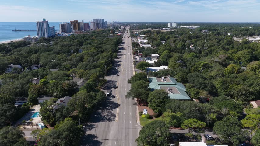 Straight road lined with oak trees runs toward the beach, a long pier and oceanfront hotel towers in Myrtle Beach, South Carolina, USA, a pool and a green roofed building beside it. Aerial view