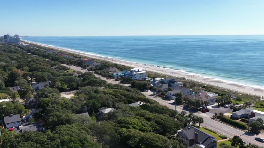 Flight follows a coastal road past large white oceanfront houses with pools and palms, a sandy beach and turquoise surf run to the horizon in Myrtle Beach, South Carolina, USA. Aerial approaching shot