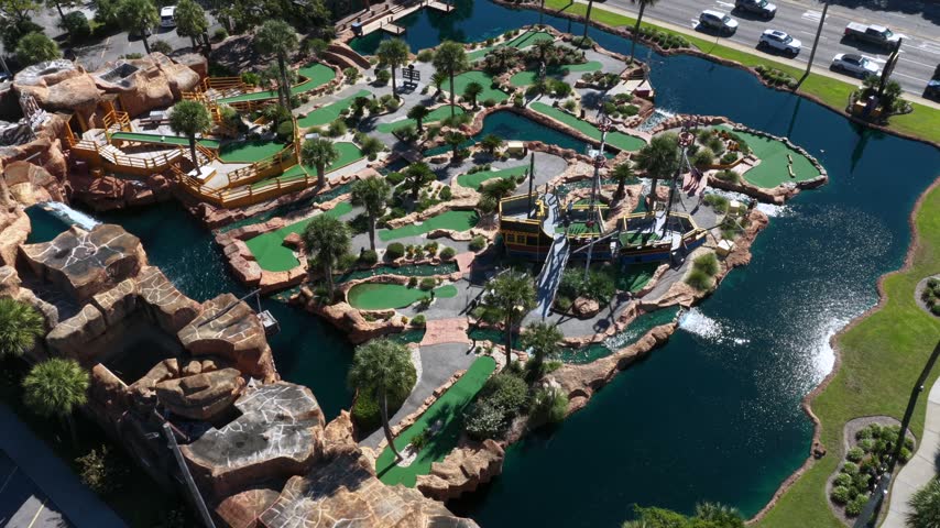 Angled view over a miniature golf course with bright green putting greens, a pirate ship, rock formations and sparkling fountains around a dark lagoon in Myrtle Beach, South Carolina, USA. Aerial view