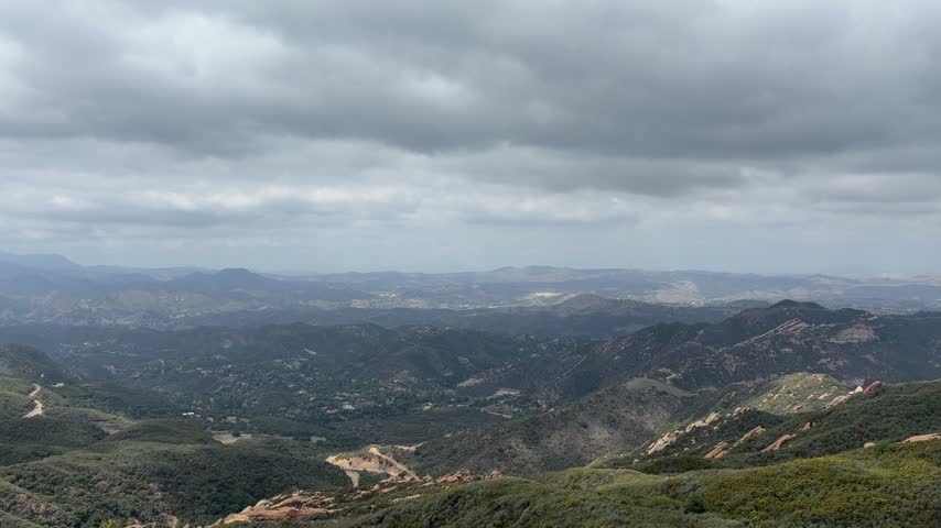 Time lapse of heavy gray clouds rolling over the canyons and red sandstone ridges of the Santa Monica Mountains near Topanga, California, with a winding road below on an overcast day.