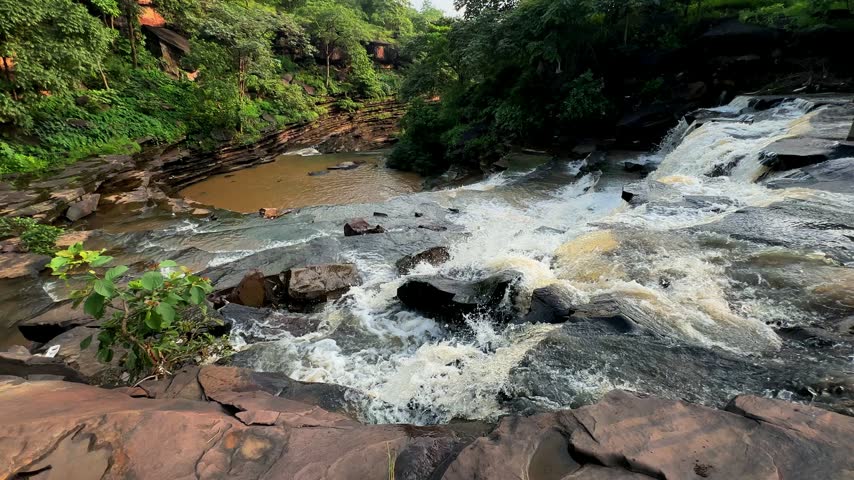Wide scenic view of cascading rapids flowing over layered rocks through dense green vegetation and a natural forest valley