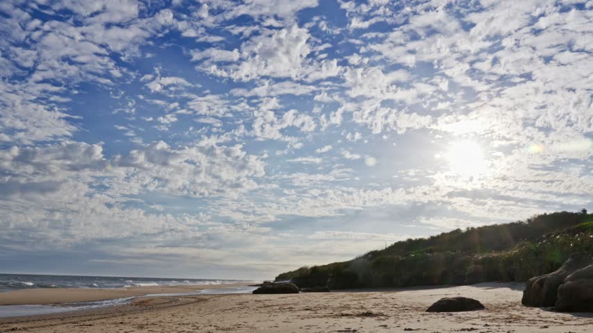 A wide angle time lapse captures the sun setting behind a vegetated dune on a sandy beach under a sky filled with dramatic clouds.