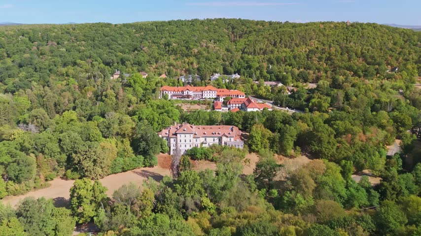 Drone aerial view of historic castle and resort buildings on a wooded hillside, surrounded by lush summer forest in Parádfürdő, Hungary, in the Mátra Mountains.