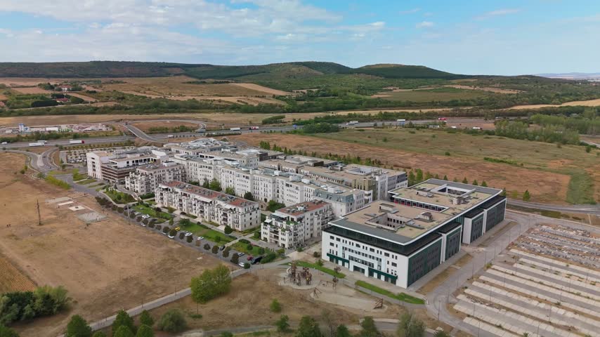 Aerial view of Tópark residential complex beside the M0 motorway in Biatorbágy, Hungary, with apartment blocks, roads, parking areas and dry farmland beneath wooded hills.