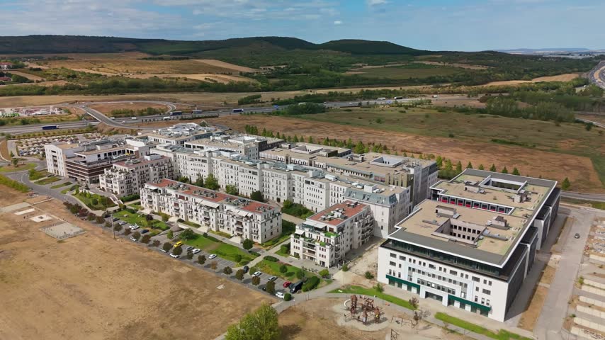 Aerial view of a modern residential complex with apartment blocks, streets and a playground beside the M0 motorway in Biatorbágy, Hungary, amid fields and wooded hills.