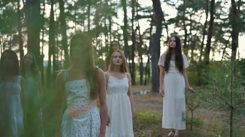 Women dressed in white gowns stand together in a forest, surrounded by trees and soft natural light, evoking a sense of sisterhood and magic