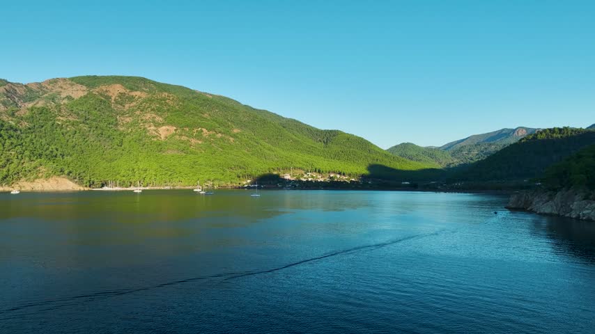 View of a calm bay surrounded by green forested hills under a clear sky with several sailboats anchored near the shore