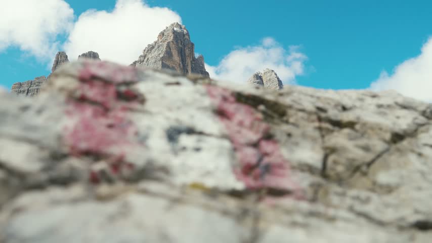 Red and white hiking trail marker painted on a rock with the iconic Tre Cime di Lavaredo peaks rising in the background in the Italian Dolomites.