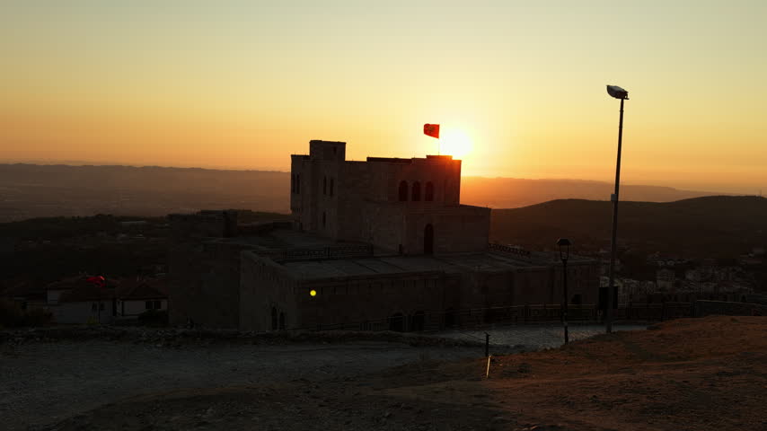 The sun sets behind the Skanderbeg Museum inside Kruje Castle, Albania. A red flag flies above the stone building, with hills in the distance.