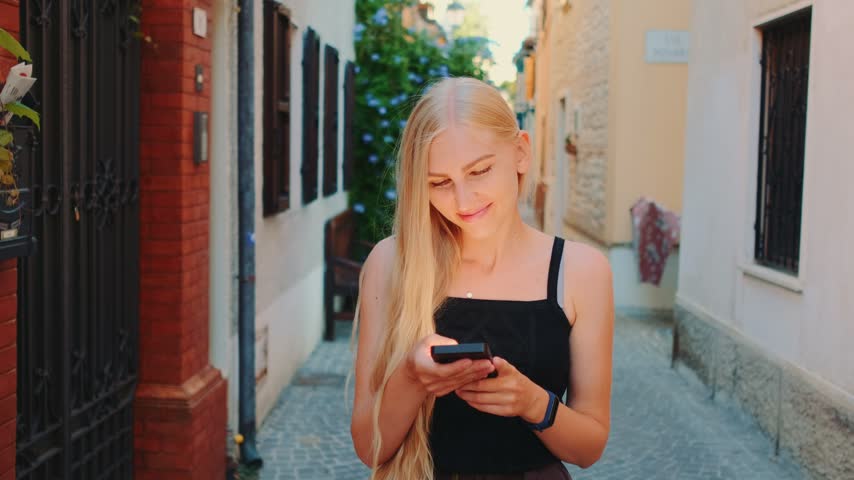 Young blonde woman wearing black tank top interacts with smartphone in a narrow cobblestone alley between brick and stucco buildings during daytime.