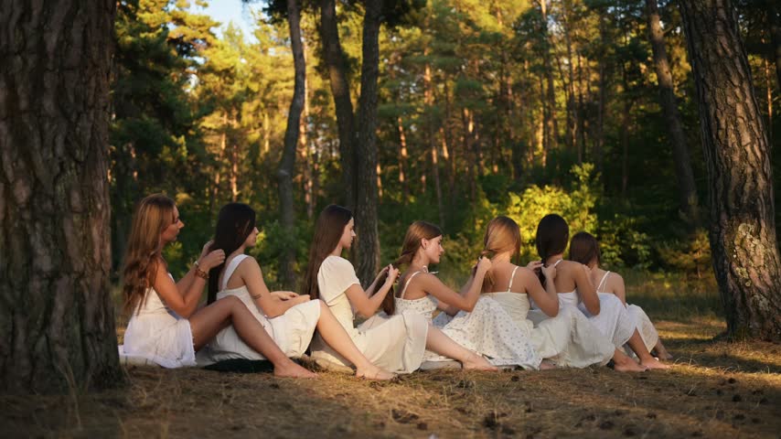 Young women in white dresses sit on the ground in a forest, braiding each other