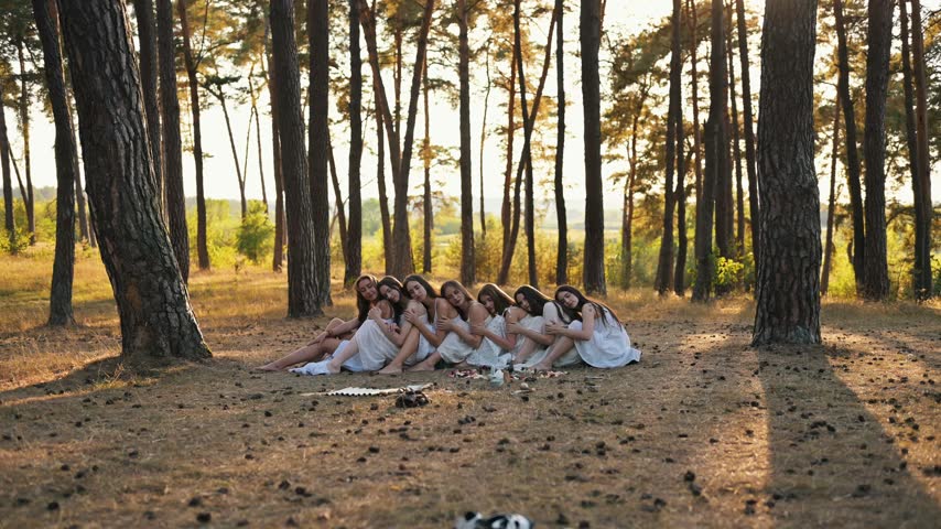 Young women dressed in white sit closely together on grass in a forest, with sunlight filtering through trees, creating a tranquil outdoor scene