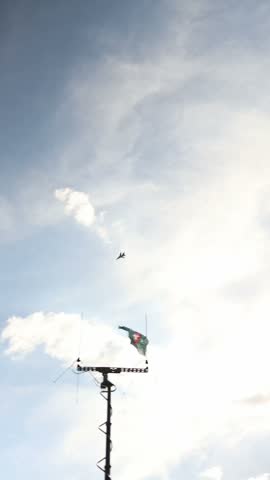 Vertical screen Military fighter jet flies across a bright cloudy sky leaving thin vapor trails above a tall communications antenna with a flag waving in the wind during daylight