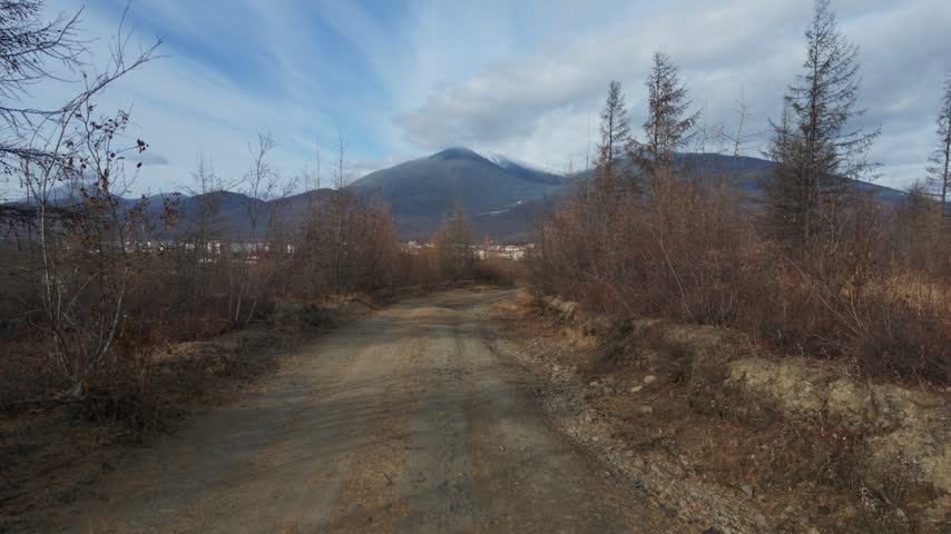 A dirt road winds through a barren, autumnal landscape with leafless trees and dry brush, leading toward a distant town at the base of snow-capped mountains under a partly cloudy sky.