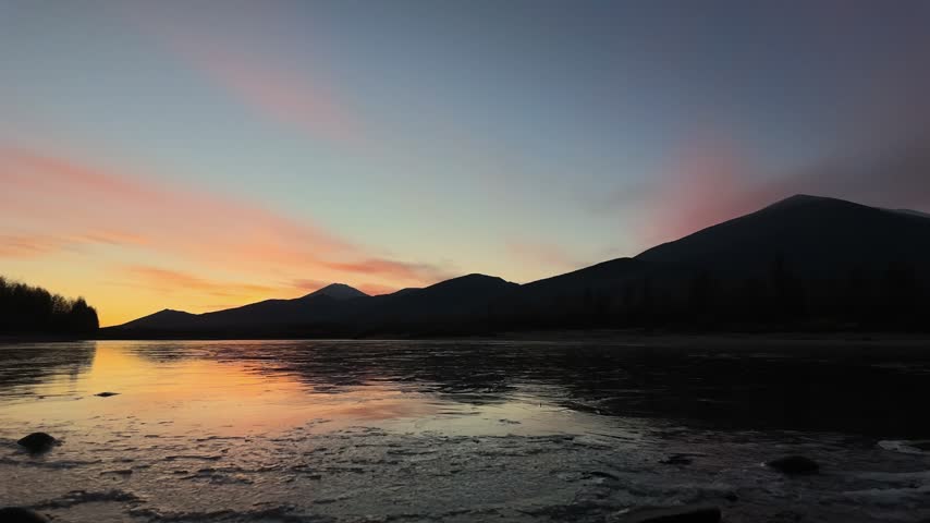 A serene sunset over a partially frozen lake reflects fading orange and pink hues as clouds drift across the horizon, silhouetting distant mountains and sparse trees.