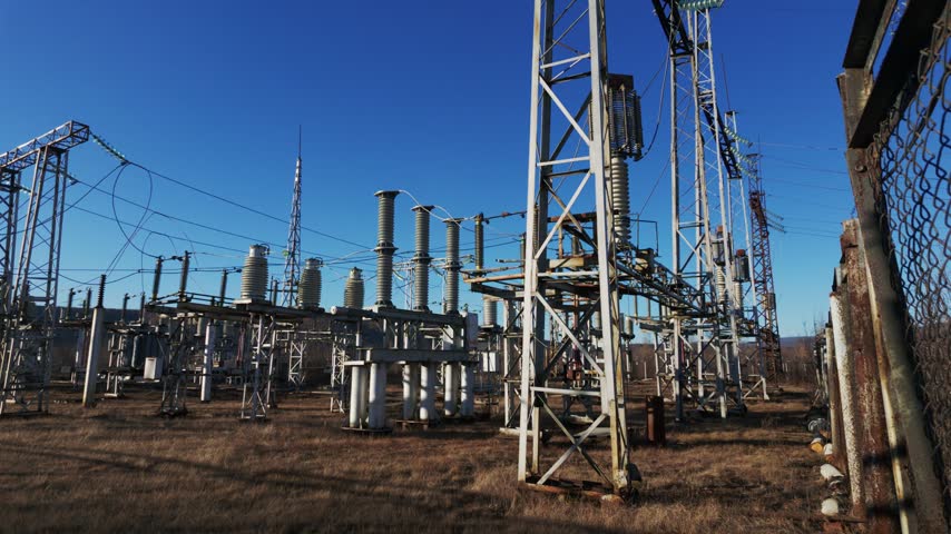 An outdoor electrical substation with metal towers, transformers, and insulators stands on dry brown grass under a clear blue sky, while shadows lengthen across the ground as the sun moves.