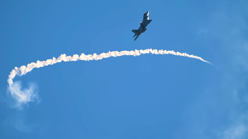 An F-22 Raptor Stealth Fighter Jet Dispensing Defensive Flares Creating Smoke Trails Across Clear Blue Sky.