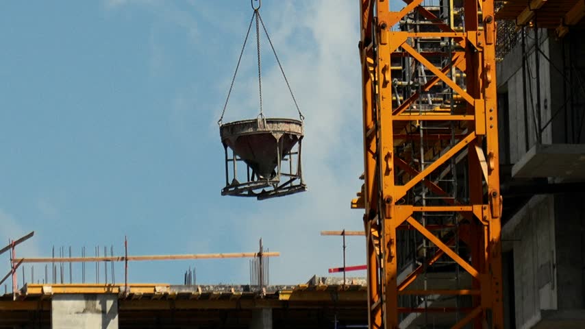 Tower crane lowers concrete bucket next to building framework under clear blue sky. Suspended container descends slowly beside tall yellow machine during highrise construction project work.