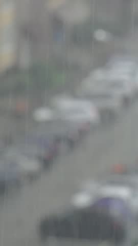 Pouring rain descends over blurred city road with vehicles parked beside walkway. Heavy rainfall creates tranquil moody ambiance amidst out of focus urban architecture on cold overcast day.