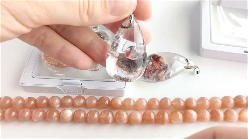 Detailed analytical examination of a transparent teardrop quartz pendant featuring distinct dark mineral inclusions, held against a backdrop of natural sunstone beads.