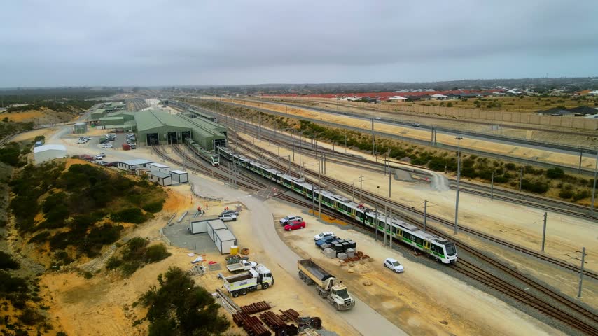 Wide view of a commuter train entering a maintenance depot facility with parked vehicles and distant residential housing on the horizon
