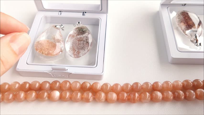 Detailed analytical examination of a transparent teardrop quartz pendant featuring distinct dark mineral inclusions, held against a backdrop of natural sunstone beads.