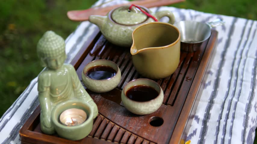 Ceramic tea set sits beside lit Buddha candle holder on slatted wooden tray outdoors. Peaceful tea arrangement encourages mindful relaxation, with cups and pitcher ready to serve. Cups accompany