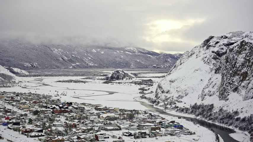 Elevated panorama of snowy El Chalten town beside a winding river with steep white cliffs and distant mountains under cloudy skies in Santa Cruz province in Argentine Patagonia