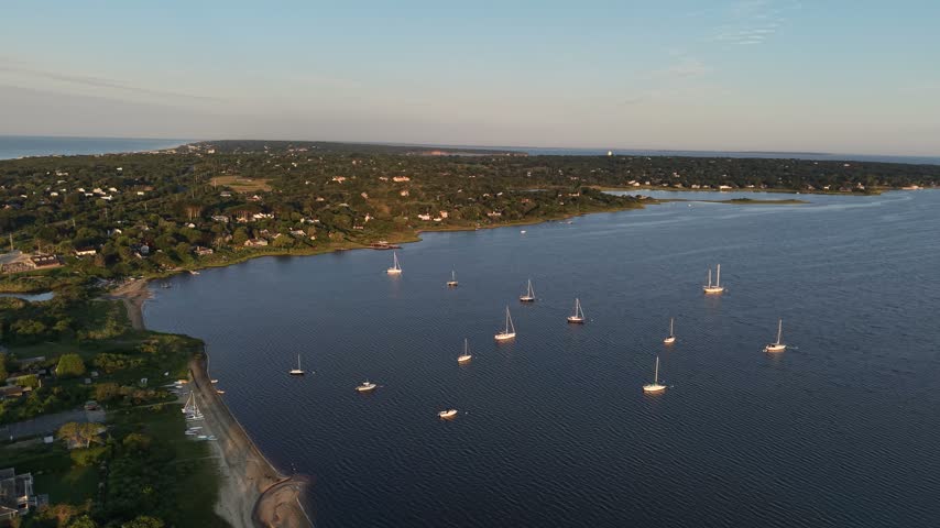Sailboats float peacefully across calm blue bay bordered by wooded peninsula and distant shoreline stretching toward horizon at sunrise in Montauk, Long Island, New York. Aerial drone wide shot