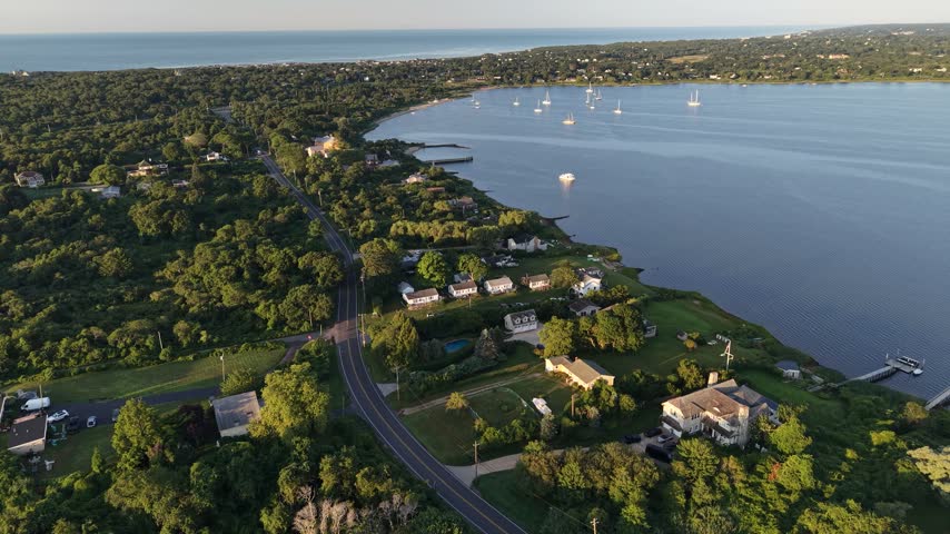 Calm bay dotted with anchored sailboats opens beside winding coastal road and waterfront homes near distant marina at golden sunrise in Montauk, Long Island, New York. Aerial drone flyover shot