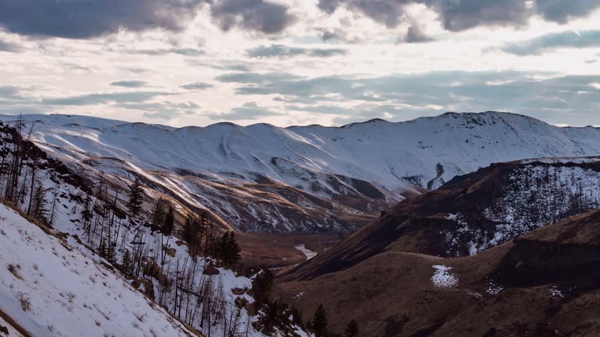 Idaho mountain range timelapse showing dramatic cloud movement over snow dusted summits and a winding river valley in a vast rural wilderness setting