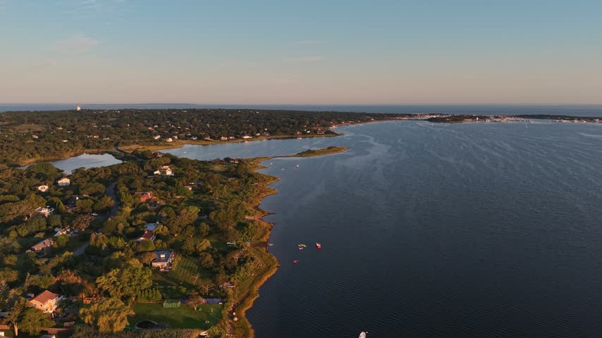 Wooded shoreline curves along calm open bay water dotted with small boats, scattered homes nestled among green trees on Long Island, New York, warm golden sunset glow. Aerial drone wide shot