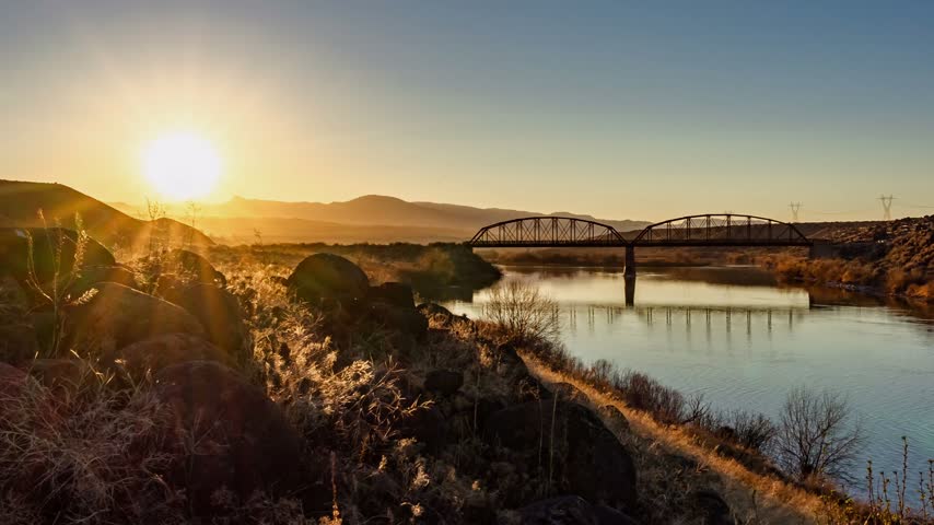 Sunset timelapse over rural Idaho river bridge featuring glowing sunlight reflecting on tranquil water and rocky shoreline with mountains