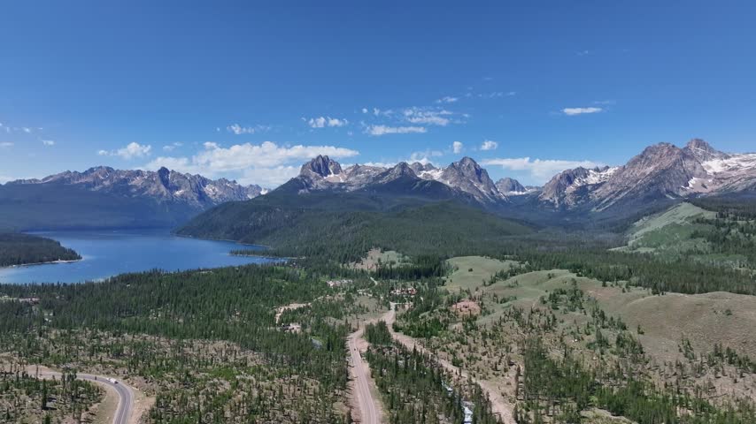 Idaho landscape unfolds from above with towering jagged peaks deep blue water and vast evergreen wilderness surrounding a rural highway