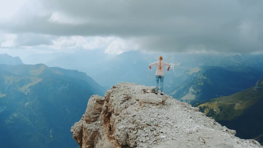 A woman stands with outstretched arms on a large rock outcrop surrounded by layered mountain ranges under an overcast sky during daytime.