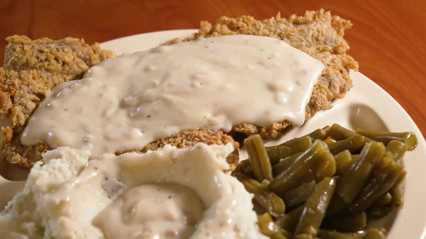 Close-up of crispy chicken fried steak topped with creamy white gravy, served with mashed potatoes and green beans on a white plate under warm restaurant lighting.